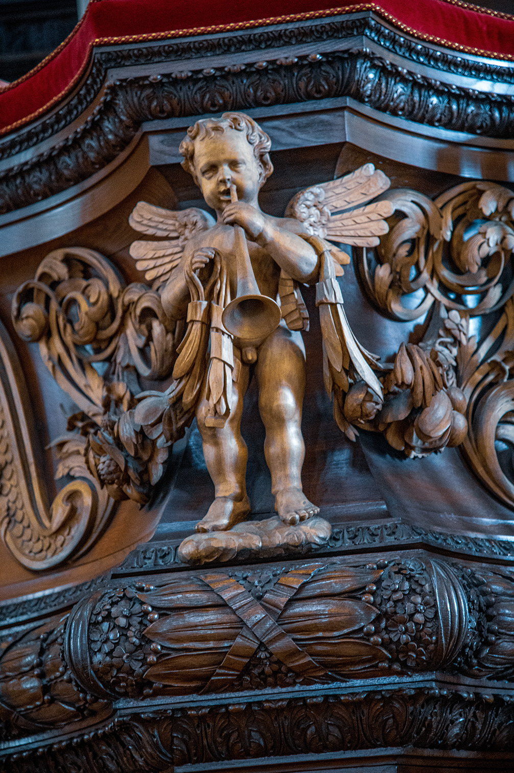The image depicts an intricately carved wooden sculpture of a cherub, or small angel, holding a trumpet. The cherub is part of a larger decorative piece, possibly an altar or pulpit, adorned with elaborate scrollwork and other ornamental details. The craftsmanship highlights fine details in the cherub's features and attire, as well as the surrounding decorations.