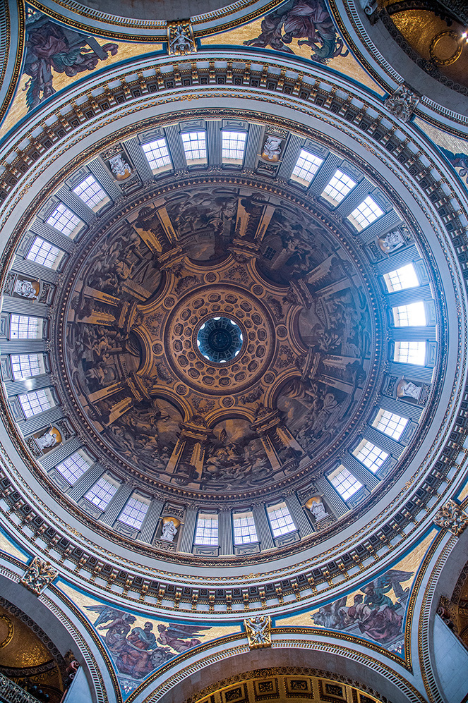 The image depicts the ornate interior dome of a grand architectural structure, likely a cathedral or a basilica. The dome is adorned with intricate frescoes, detailed carvings, and statues. It features a series of windows that allow natural light to illuminate the space, highlighting the elaborate artwork and architectural details.