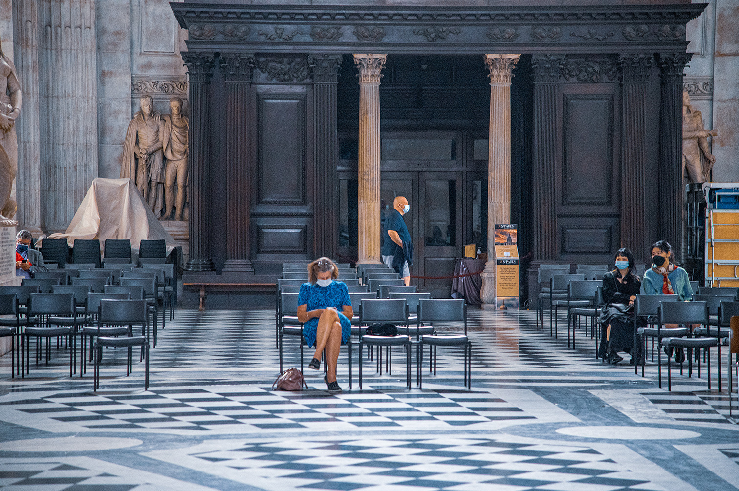 The image depicts an interior of a large, ornate building, likely a church or cathedral, with high ceilings and grand architectural elements such as columns and statues. Several people are seated in chairs, spaced apart, and all are wearing face masks, indicating adherence to social distancing guidelines. The floor is covered with a checkered black and white marble pattern. The scene suggests a quiet, possibly pandemic-era setting where people are waiting or resting.