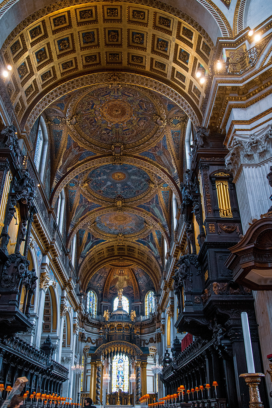 The image depicts the interior of a grand, ornate cathedral. The ceiling is adorned with intricate gold patterns and detailed frescoes. The architecture features tall, elaborately decorated columns and arches. Stained glass windows and chandeliers add to the opulence. The overall atmosphere is one of historical and artistic significance, showcasing the grandeur of religious architecture.