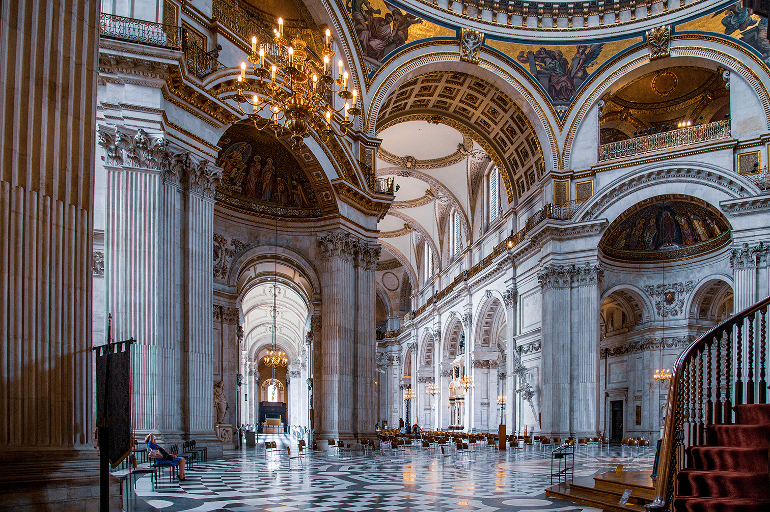 The image depicts the interior of a grand, ornate cathedral or church. The architectural style is classical with tall, white marble columns, intricate carvings, and detailed frescoes adorning the ceilings and domes. The space is illuminated by natural light streaming through large windows and by several large chandeliers hanging from the ceiling. The floor is covered with a patterned, polished stone. The overall atmosphere is one of grandeur and reverence, typical of significant historical and religious buildings