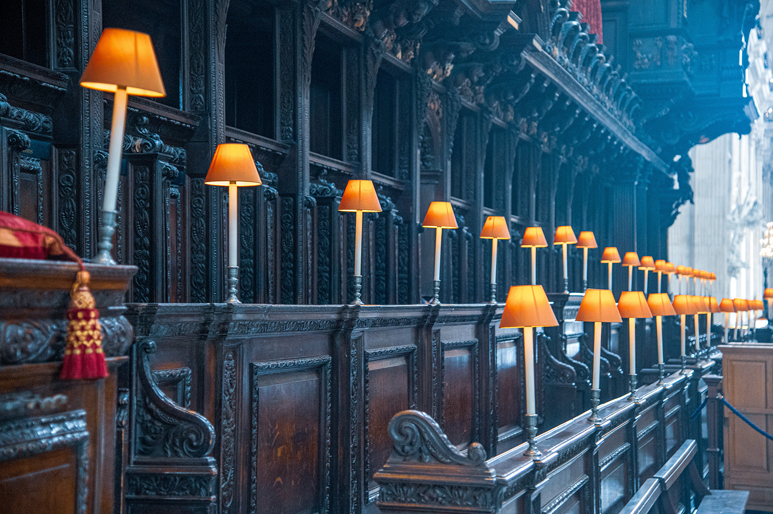 The image depicts an ornate, dark wooden interior with a series of lamps with yellow shades lining the top of the pews, likely in a church or cathedral. The intricate carvings on the wood and the detailed design elements contribute to a sense of grandeur and historical significance.