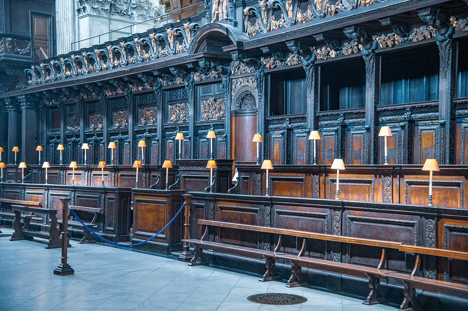 The image depicts an ornate, historical interior, likely a courtroom or a council chamber. The room features intricate wooden paneling with detailed carvings and sculptures. Rows of wooden benches with reading lamps are arranged neatly, and the area is cordoned off with blue ropes and stanchions. The architecture includes elaborate moldings and statues, suggesting a significant and grand setting.