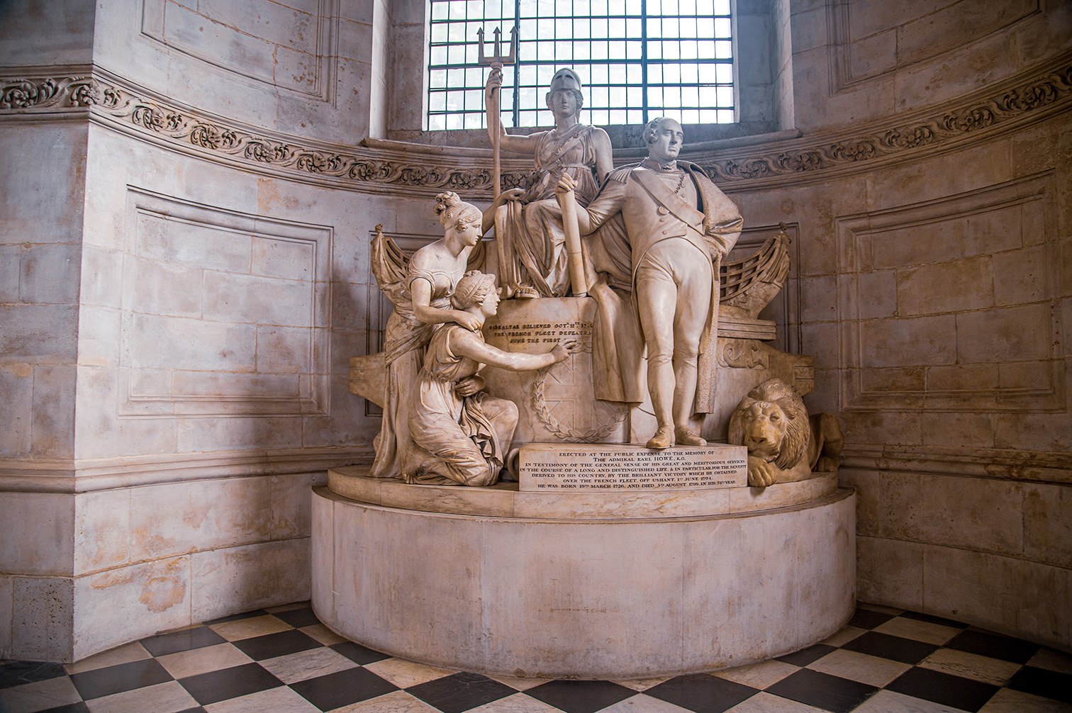 The image depicts a marble statue memorial in a grand, classical interior setting. The statue features a prominent figure in naval uniform standing beside a seated female figure, likely representing a personification of Britain or Victory. Another female figure is kneeling at the base, and a lion is lying at the forefront. The memorial is inscribed with text commemorating Admiral Earl Howe, detailing his significant naval victories and contributions, particularly the defeat of the French fleet off Ushant on 1 June 1794. The memorial was erected at public expense and stands as a testament to his long and distinguished life and meritorious services.