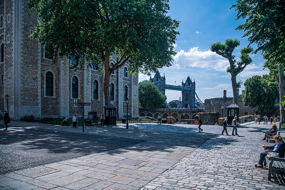 The image depicts an outdoor scene at the Tower of London, with the Tower Bridge visible in the background. The area is paved with cobblestones and features historic stone buildings with arched windows. There are several people walking and sitting, enjoying the sunny day. Trees provide some shade, and the sky is clear with a few clouds. The overall atmosphere is calm and leisurely.