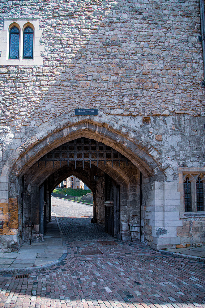 A sombre sign, etched with the fateful words &ldquo;BLOODY TOWER&rdquo;, hangs like a pronouncement above a formidable archway. This is no gentle embrace, but a stern invitation into the heart of ancient mysteries. The arch, a masterpiece of medieval engineering, is guarded by a fearsome portcullis, its iron teeth poised as if ready to descend. It speaks of security, yes, but also of confinement, of secrets held tight within these hallowed walls.