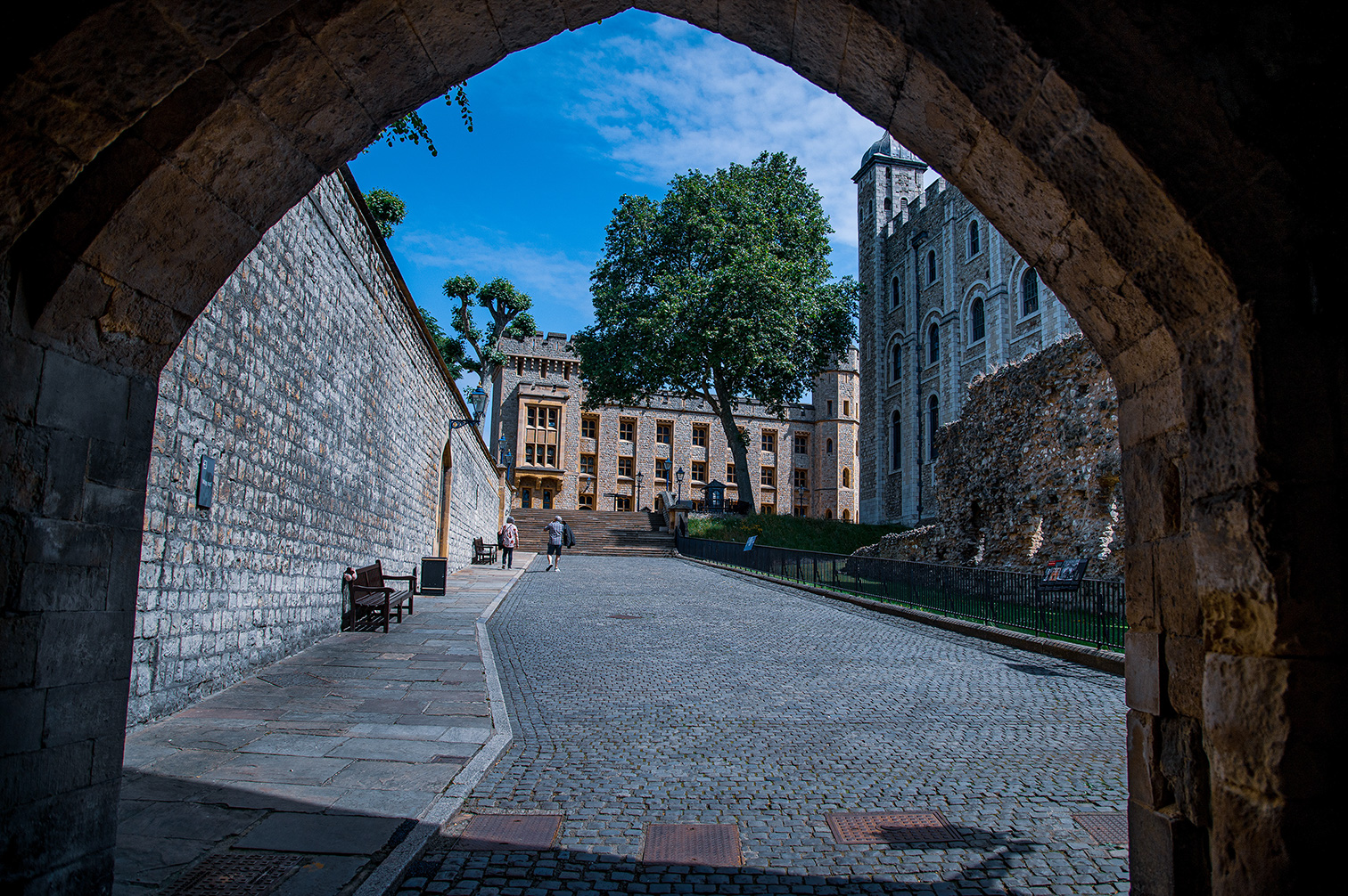 The image depicts a view through an archway of a historical stone structure, likely a castle or fortress. The scene includes cobblestone pathways, stone walls, and a large, tall building with a tower. There are a few people walking and benches along the path. The sky is clear with some clouds, and trees are visible in the background.
