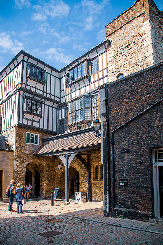 The image depicts a historic building with a distinctive Tudor architectural style, featuring black and white timber framing. The structure includes multiple stories with large windows and a prominent entrance archway. The building is constructed with a combination of stone and brick, and the scene is set on a cobblestone street. A few people are seen walking near the entrance, and the sky above is clear with some clouds. The overall atmosphere suggests a well-preserved or restored historic site, likely in a European setting.