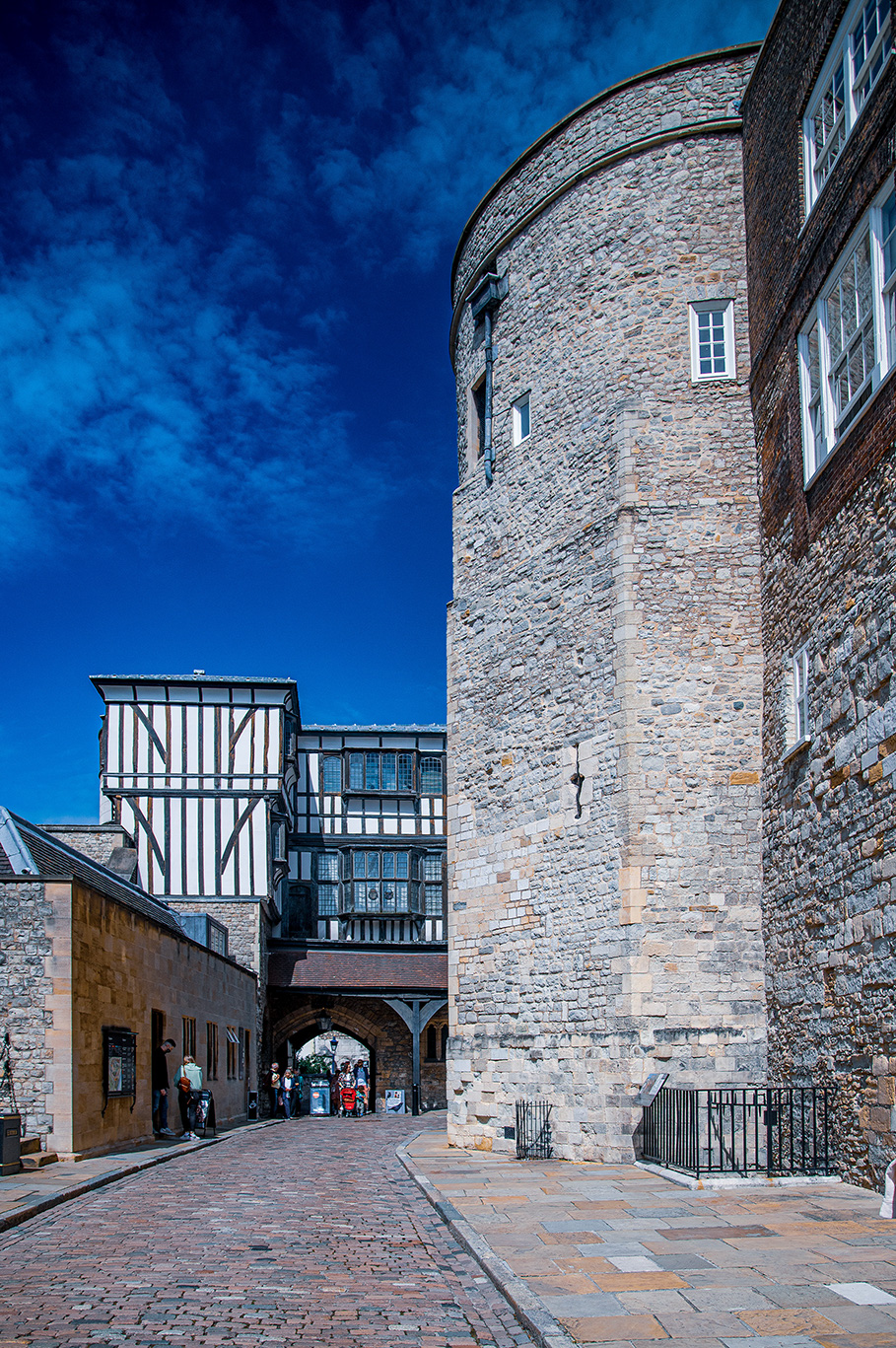 The image depicts a historic architectural setting with a cobblestone path leading to a large, round stone tower on the right. The tower features small windows and a metal lantern attached to its wall. Adjacent to the tower is a half-timbered building with distinctive black and white wooden beams. Several people are seen walking or standing along the path, and the sky above is clear and blue.