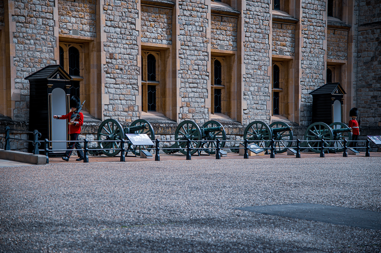 The image depicts a scene in front of a historic stone building, likely a castle or fortress, with two guards in traditional red uniforms and bearskin hats standing at attention. In front of the guards, there are several old cannons displayed on a gravel area. The building features large stone blocks and tall, narrow windows, contributing to its historical and majestic appearance.