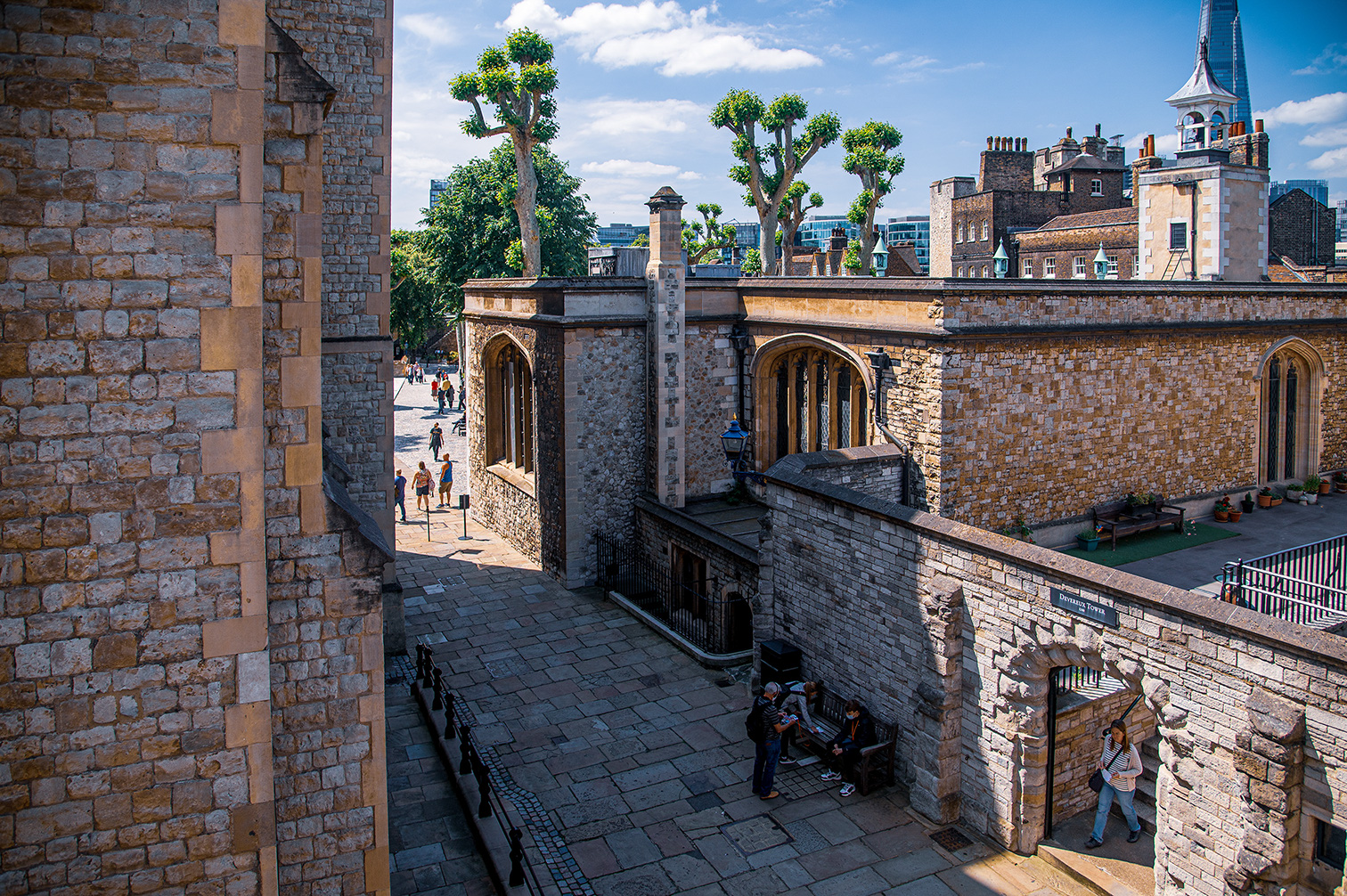 The image depicts a historic stone structure, likely a castle or fortress, with people walking and sitting in the courtyard. The architecture features arched doorways and windows, and the walls are made of large, weathered stones. The scene is bathed in sunlight, and modern buildings can be seen in the background, indicating an urban setting. The overall atmosphere is one of historical significance blended with contemporary life.