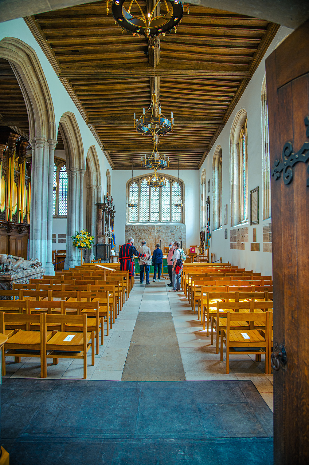 The image shows the interior of a church with rows of wooden pews and a group of people standing at the front. A person in traditional attire is addressing the visitors.
