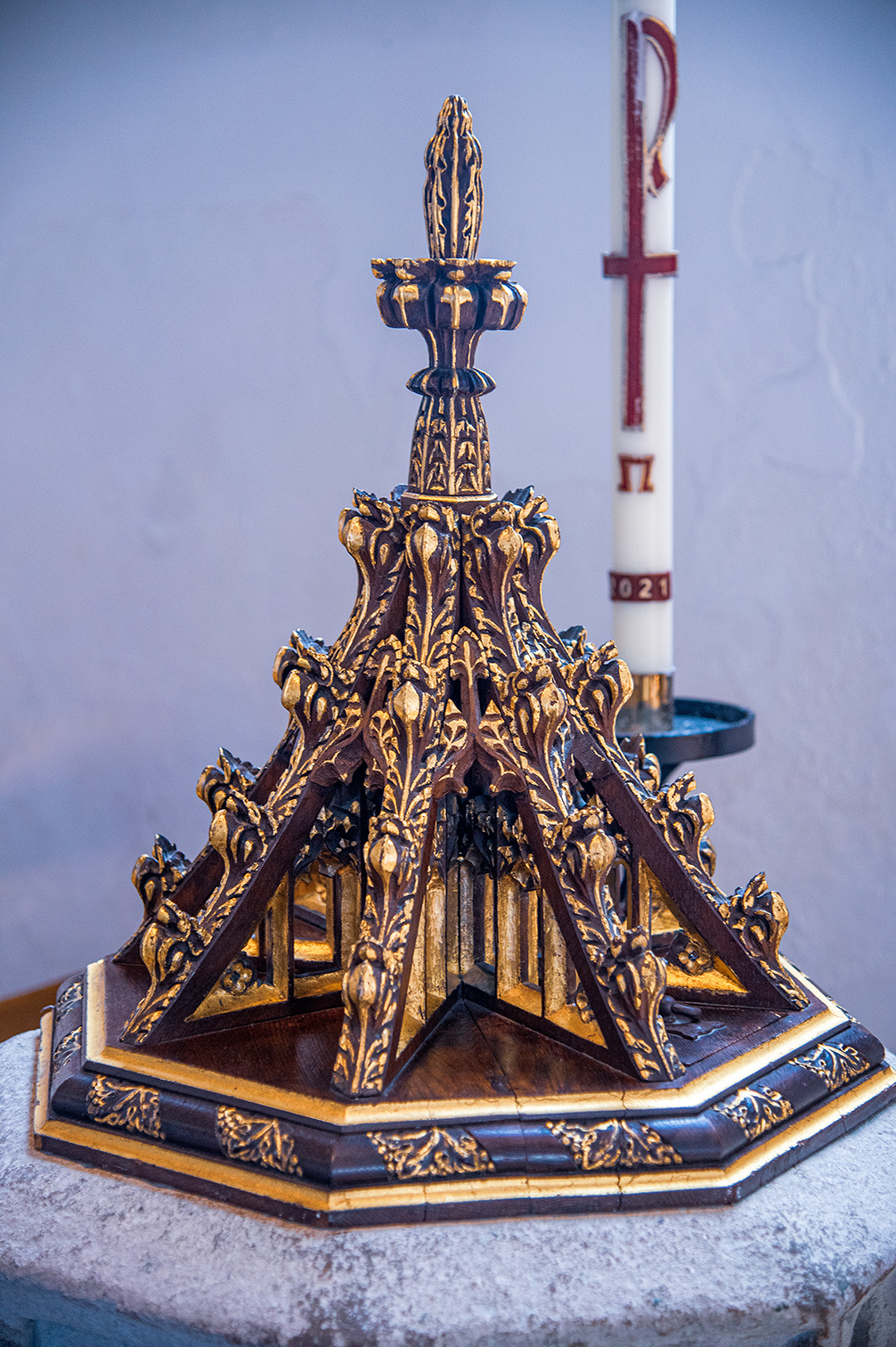 A highly ornate, dark wood and gold decorative piece sits on a stone surface. To the right of the piece is a tall white candle with red religious markings.