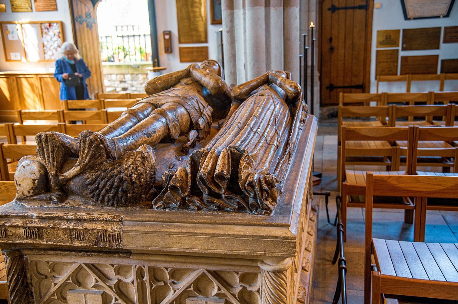 This image depicts a tomb with effigies of a man and a woman, likely a knight and his lady, lying side-by-side. The effigies are detailed, with the man depicted in armor and the woman in a flowing gown. They are placed on a stone sarcophagus adorned with intricate carvings. In the background, we see wooden pews typical of a church or chapel setting, and a woman in a blue jacket looking at a notice board. The overall ambiance suggests a historical and solemn location within a religious building.