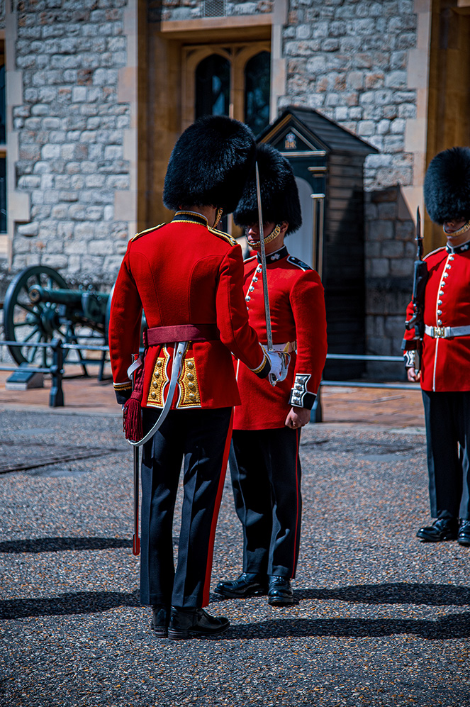 Three guards in red ceremonial uniforms with tall black furry hats stand at attention on a paved surface. They are holding swords and appear to be guarding a building with a stone facade.