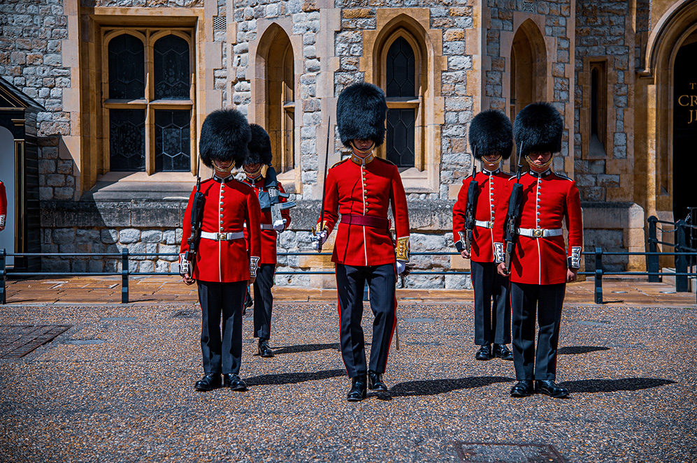 The image depicts a group of guards dressed in traditional red uniforms and black bearskin hats, marching in front of a historic stone building. The guards are walking in a straight line, with the lead guard in the center and others following behind. The setting appears to be a formal or ceremonial location, likely a significant landmark or palace.