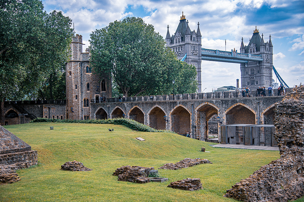 The image depicts a historical site with ancient ruins in the foreground and a modern bridge in the background. The ruins are surrounded by a well-maintained grassy area, and there is a stone structure with arches. The bridge, known as Tower Bridge, is an iconic landmark in London, featuring two tall towers connected by a walkway. The scene is set on a clear day with blue skies and some clouds, and there are people visible on the bridge.