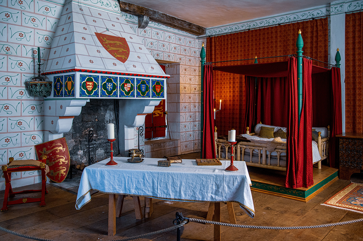 The image depicts a historical room, likely from a medieval period, featuring intricate tile work on the walls with a prominent shield emblem above a fireplace. The room includes a four-poster bed with red curtains, a table with a white cloth and various items on it, and a chair with a shield leaning against it. The overall decor suggests a setting of nobility or high status.