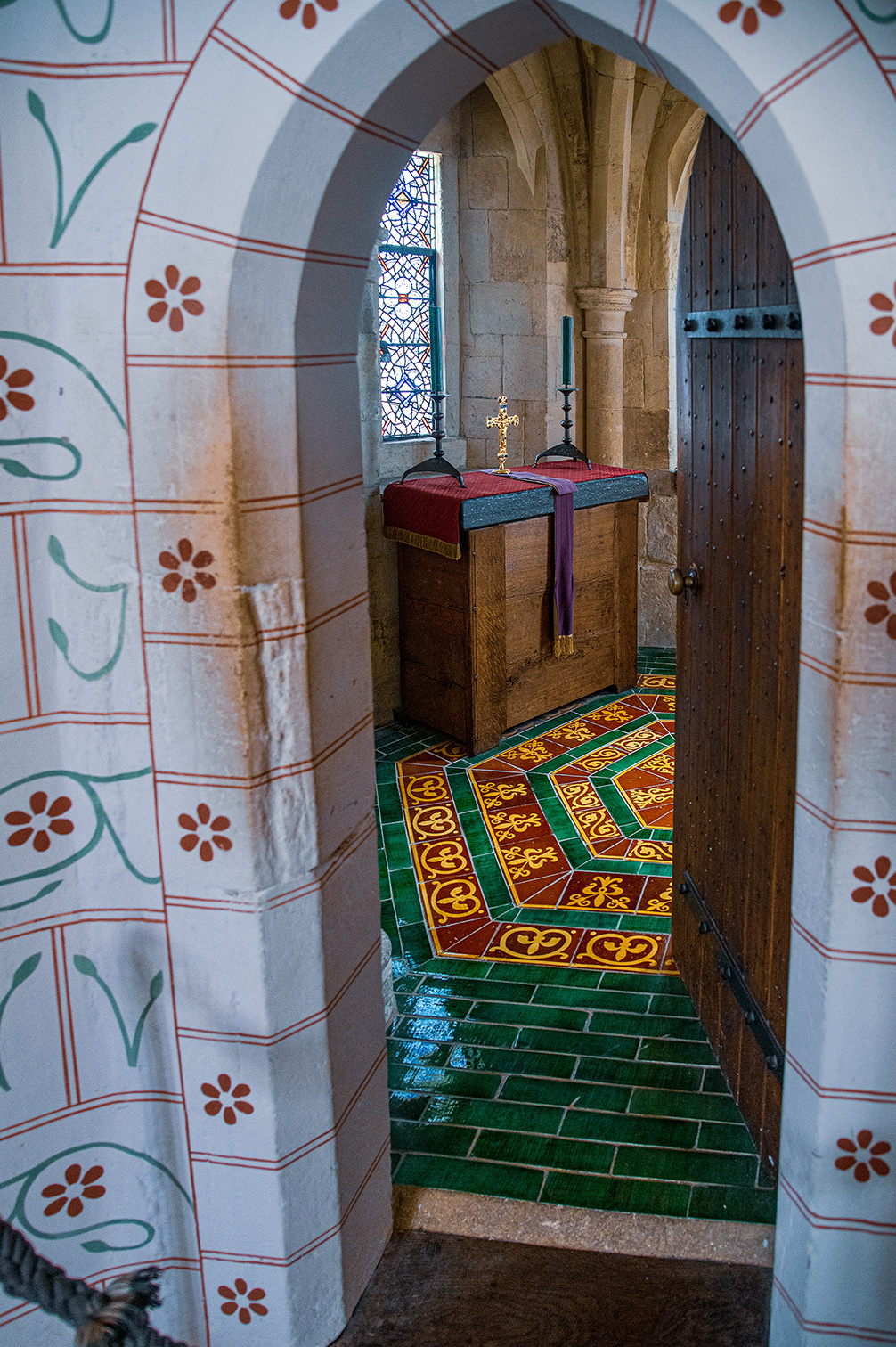 The image depicts an interior view of a historical or religious building, featuring a narrow passage with ornate tile flooring and decorated walls. The passage leads to a small altar adorned with a cross and covered with a cloth. The architecture includes arched doorways and stone columns, with a stained glass window visible in the background.