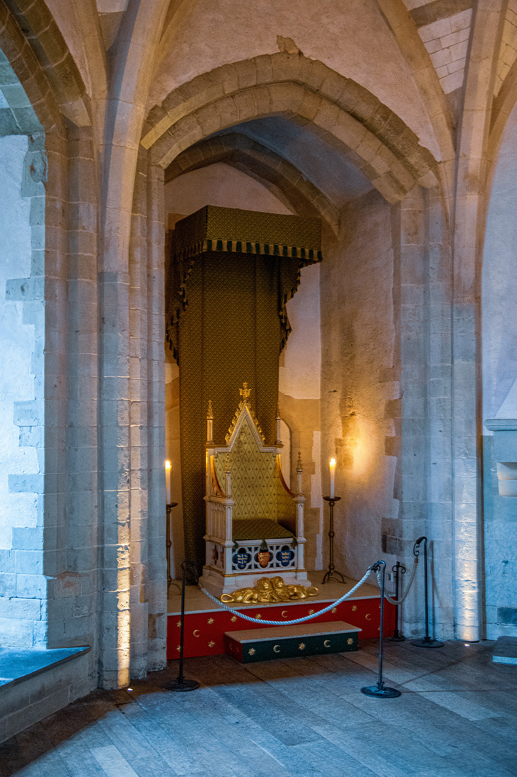 The image depicts an ornate, historical chapel or shrine, featuring a richly decorated altar with gold and intricate patterns. The setting includes stone arches and columns, with candles providing warm lighting. The area is cordoned off with ropes and stanchions, indicating it is a preserved or protected site, likely of significant cultural or religious importance.