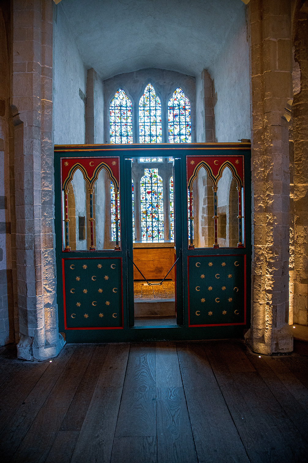 The image depicts an interior view of a historic building, likely a church or cathedral. The focal point is a wooden structure with ornate, green doors featuring gold crescents and stars, which appear to lead to a small, enclosed area. Behind the structure, there are three tall, arched stained-glass windows that allow colorful light to filter into the space. The floor is made of wooden planks, and the walls are constructed of large stone blocks, adding to the ancient and reverent atmosphere of the setting.