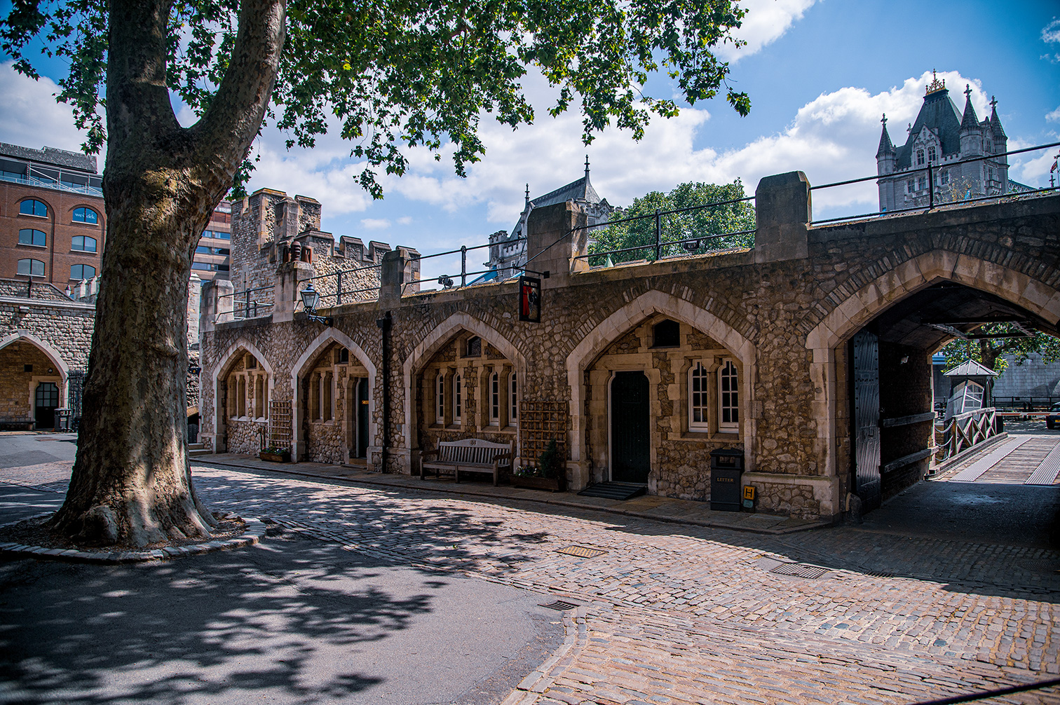 The image depicts a historic stone bridge with multiple arches and towers, likely part of an old castle or fortification. The structure features Gothic architectural elements such as pointed arches and detailed stonework. The bridge spans over a road with cobblestone paving, and there is a tree on the left side of the image casting shadows on the ground. The sky is partly cloudy, and modern buildings are visible in the background, indicating an urban setting blending historical and contemporary elements.