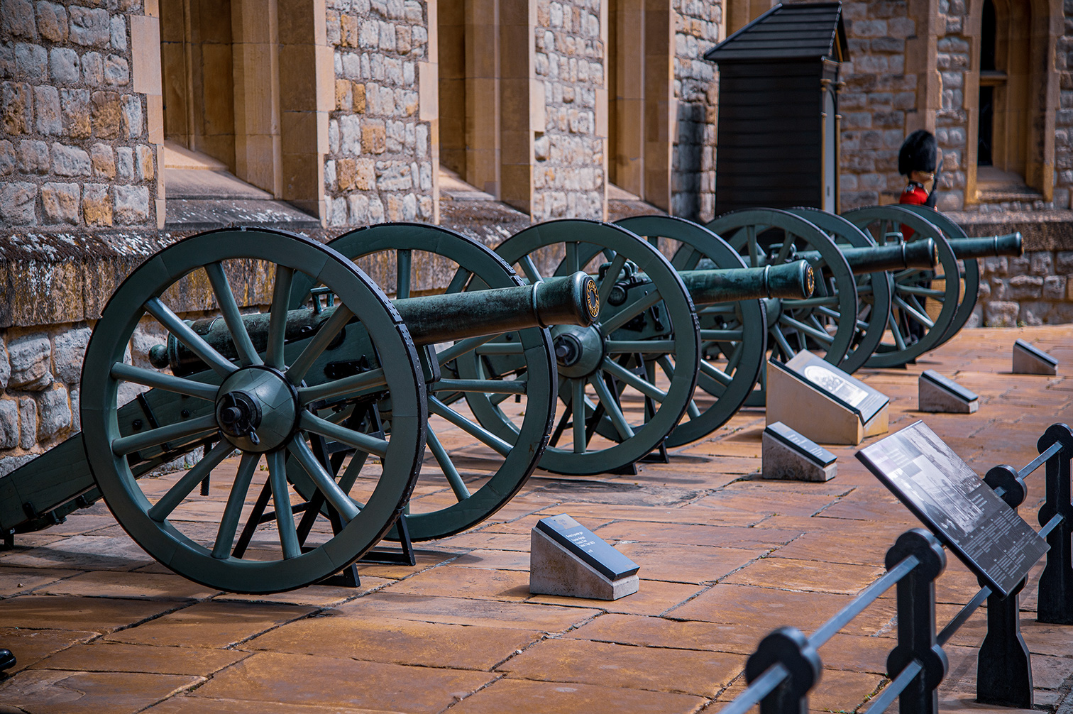 The image depicts a row of historical cannons displayed outdoors, likely in a museum or historical site setting. The cannons are mounted on large wheels and are positioned in front of a stone building with arched windows. Informational plaques are placed in front of each cannon, providing details about them. The scene includes a guard in traditional attire standing near the building, adding to the historical ambiance.