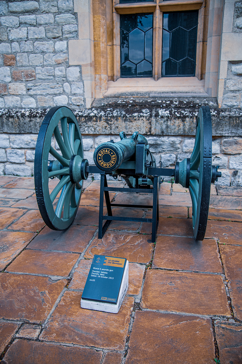 The image shows an old bronze cannon mounted on wooden wheels, positioned in front of a stone building with a window above. The cannon has the inscription 'ROYAL ARMOURIES' on its barrel. In front of the cannon, there is an informational plaque providing details about the cannon, including its type, origin, and the date it was founded.