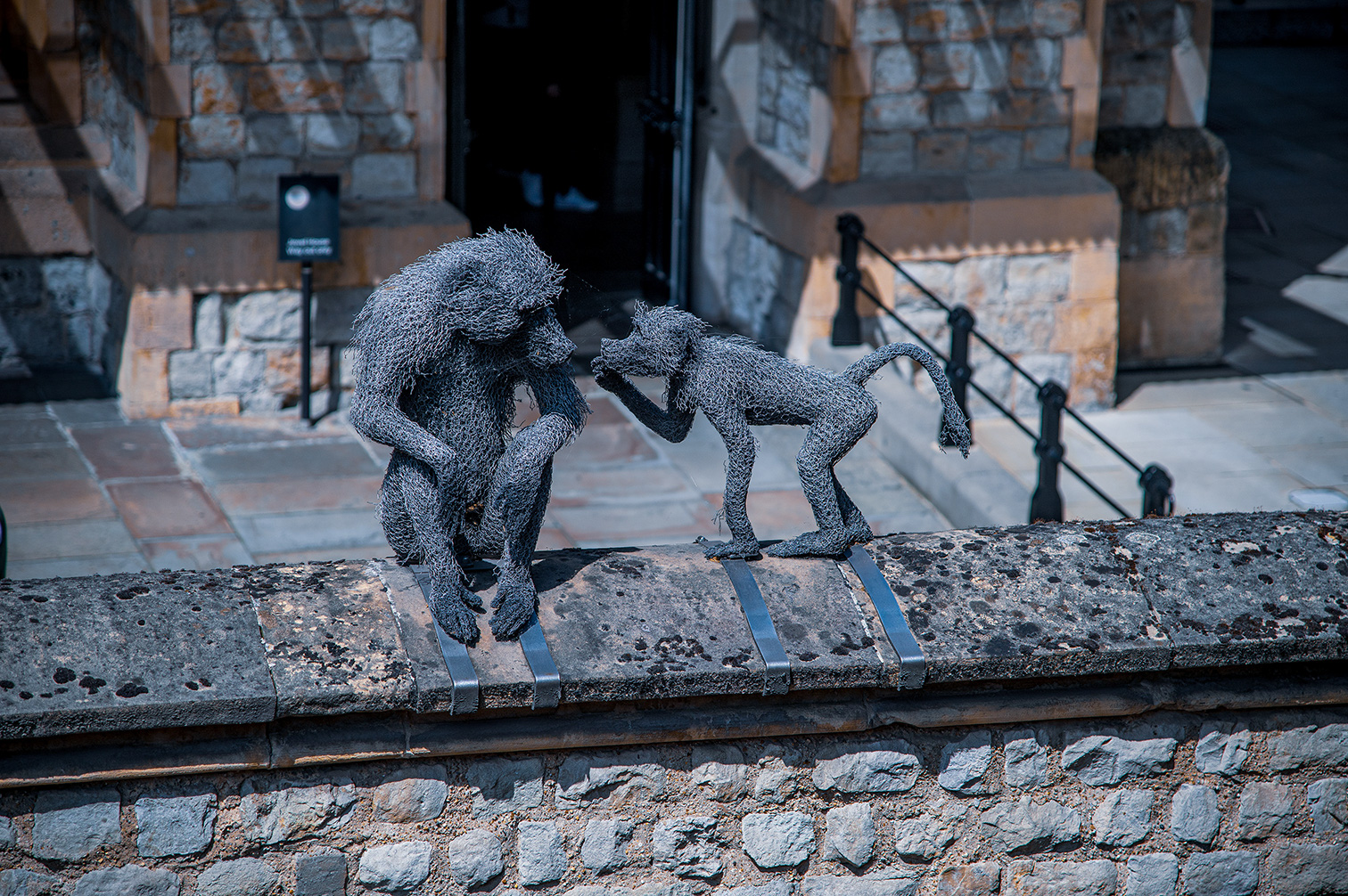 The image depicts two wire mesh sculptures of monkeys on a stone structure. One monkey is in a crouching position, while the other is standing on two legs, appearing to interact or communicate with the crouching monkey. The background features a stone wall with a wooden door, and there is a rope barrier behind the sculptures.