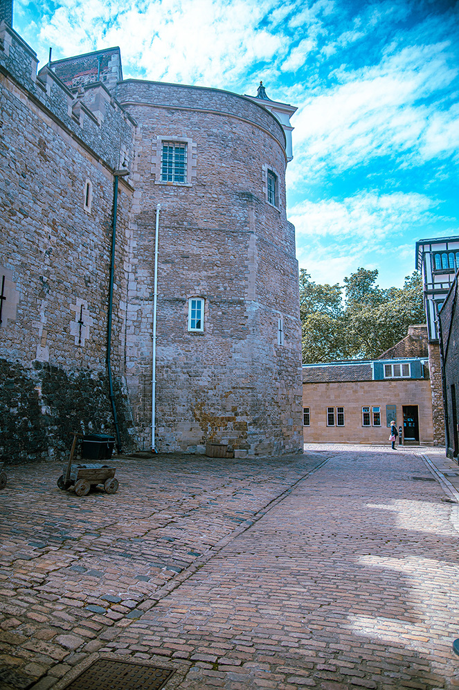The image depicts a historic stone building with a cylindrical tower, featuring small windows and a cobblestone courtyard. The sky is partly cloudy, and there is a wooden cart on the left side of the courtyard. The architecture suggests an old, possibly medieval structure.