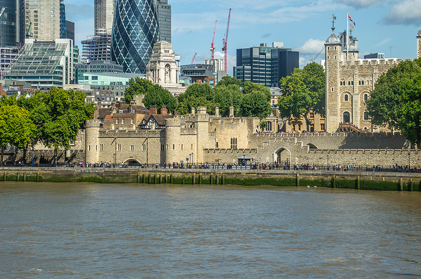 The image depicts the Tower of London, a historic castle located on the north bank of the River Thames in central London. The castle is surrounded by modern skyscrapers, showcasing a blend of historical and contemporary architecture. The scene includes the castle's stone walls, towers, and lush green trees, with people walking along the riverside path. The River Thames flows calmly in the foreground, reflecting the cityscape.
