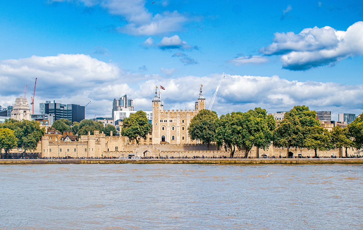 The image depicts the Tower of London, a historic castle located on the north bank of the River Thames in central London. The photograph captures the castle's distinctive architecture, including its towers and stone walls, surrounded by lush green trees. In the background, modern skyscrapers and construction cranes indicate the blend of historic and contemporary elements in the cityscape. The sky is mostly clear with some scattered clouds, and the river in the foreground adds to the scenic view.