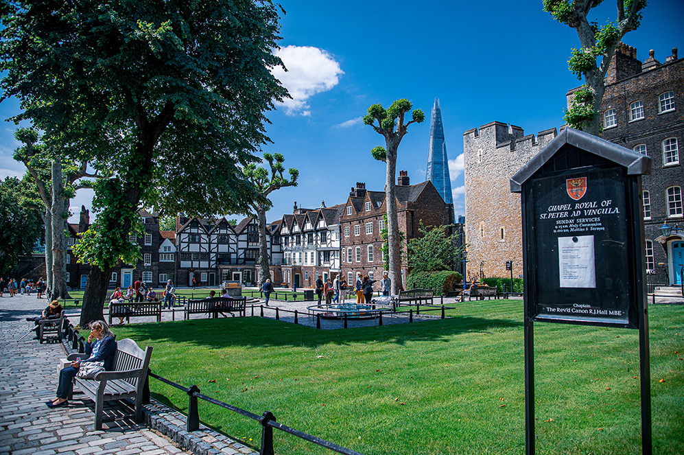 The image depicts a serene park setting with people relaxing on benches and walking around. In the background, there are historic buildings and a prominent modern skyscraper. A signboard provides information about the Chapel Royal of St. Peter ad Vincula, including details about Sunday services.
