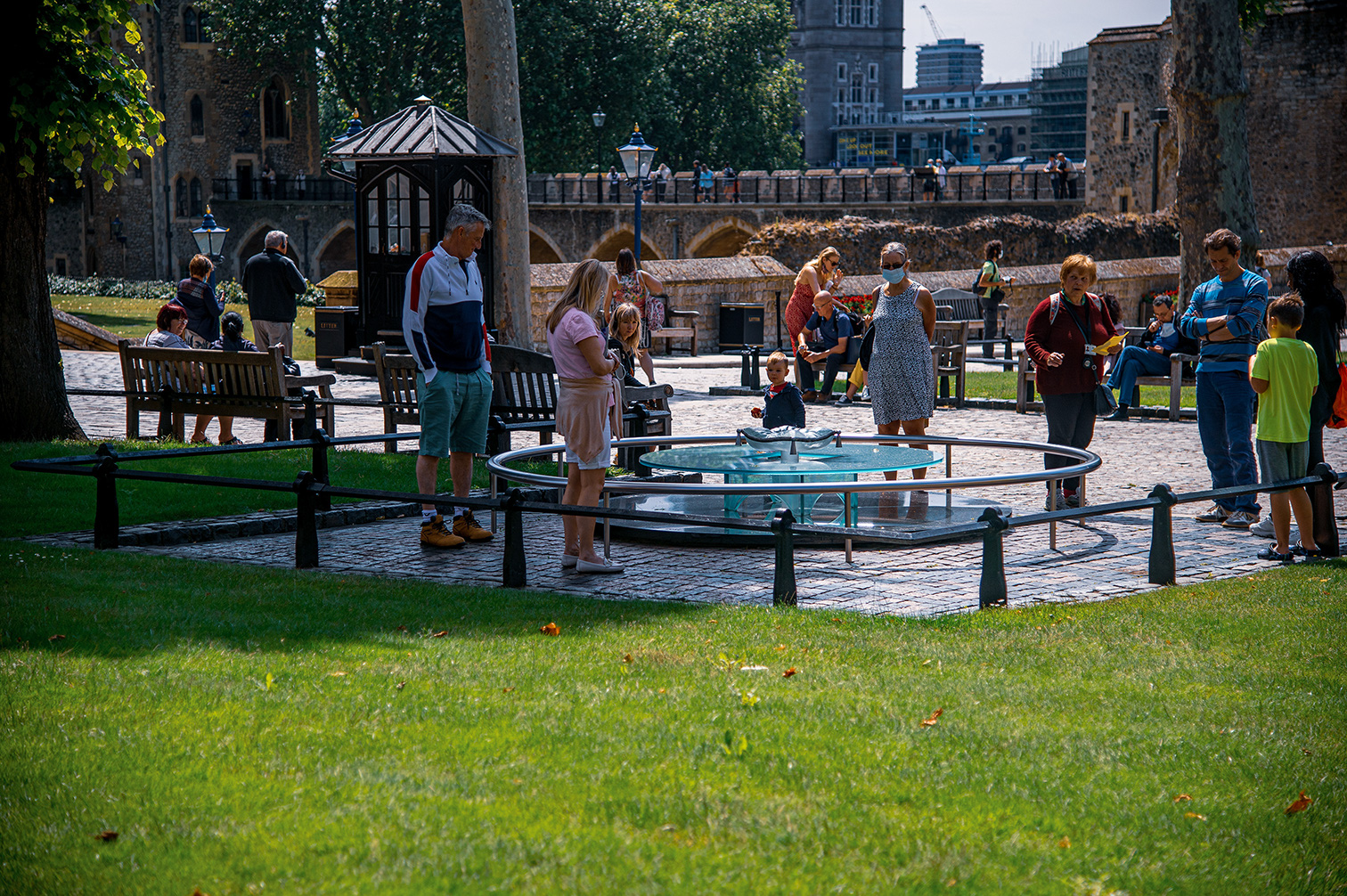 The image depicts a lively scene in a park-like setting, likely within a historic or tourist area. People of various ages are gathered around a circular fountain, engaging in conversations and observing the surroundings. The background features old stone buildings and a guard tower, suggesting a location of historical significance. The atmosphere appears relaxed and family-friendly, with individuals sitting on benches, walking around, and enjoying the day. The overall mood is calm and leisurely, with people taking time to appreciate their environment.