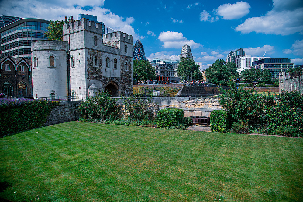 The image depicts a historic castle with modern buildings in the background, showcasing a blend of old and new architecture. The foreground features a well-maintained lawn and garden area, with a clear blue sky and scattered clouds above.