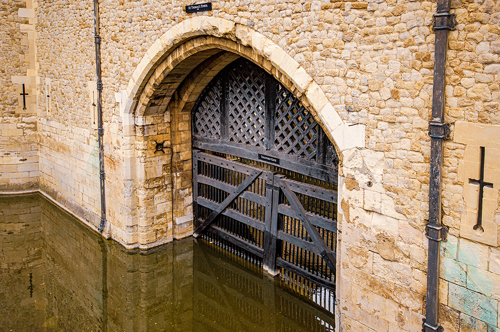 The image depicts an old stone archway with a wooden gate leading to a water-filled passage. The structure appears to be part of a historical or medieval building, featuring intricate stonework and a cross symbol on the wall. The water level is high, nearly reaching the bottom of the gate.