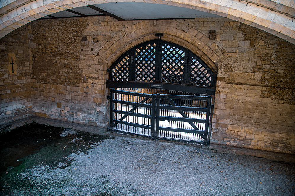 The image depicts an arched entrance made of stone bricks with a metal gate. The gate is designed with a combination of diamond and rectangular patterns. The ground in front of the gate appears to be wet, possibly due to a small puddle of water. The structure has a historical or industrial appearance, with a cross symbol visible on the left wall. The gate is labeled with the text 'Tower Gate'.