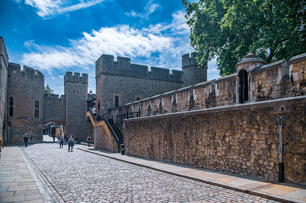 The image depicts a historic castle with stone walls and towers, likely a medieval fortress. The scene includes a cobblestone path leading up to the castle entrance, with a few people walking along the path. The sky is clear with some clouds, and there are trees visible on the right side of the image. The castle features battlements and a prominent gatehouse, indicating its defensive purpose.