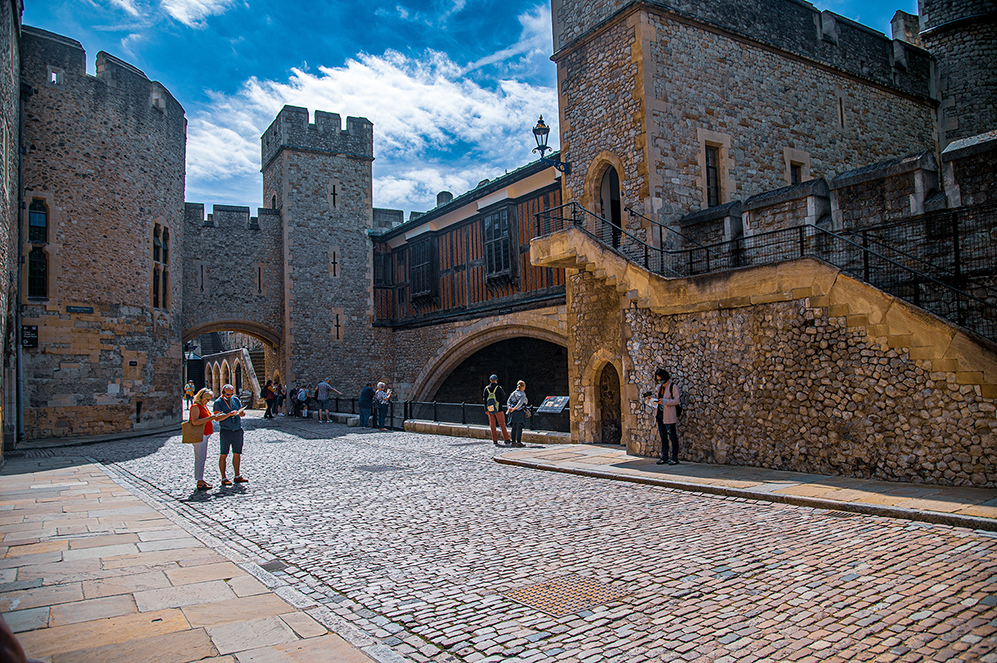 The image depicts a historic castle courtyard with stone buildings, towers, and cobblestone paths. Several people are seen walking and standing around, possibly tourists exploring the site. The architecture features arched doorways, wooden-framed windows, and a mix of stone textures. The sky is clear with some clouds, suggesting a sunny day. The overall atmosphere is that of a well-preserved historical site, likely a popular tourist destination.
