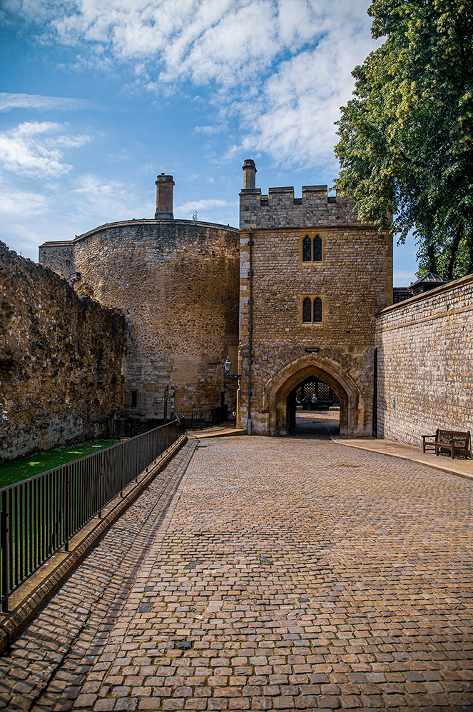 The image depicts a historic stone structure, likely part of a castle or fortress, featuring a cobblestone pathway leading to an arched gateway. The architecture includes a round tower on the left and a square tower with battlements on the right. The sky is partly cloudy, and there is greenery visible on the right side of the image. The pathway is bordered by a metal fence on the left and a stone wall on the right, with a bench visible on the right side near the wall.