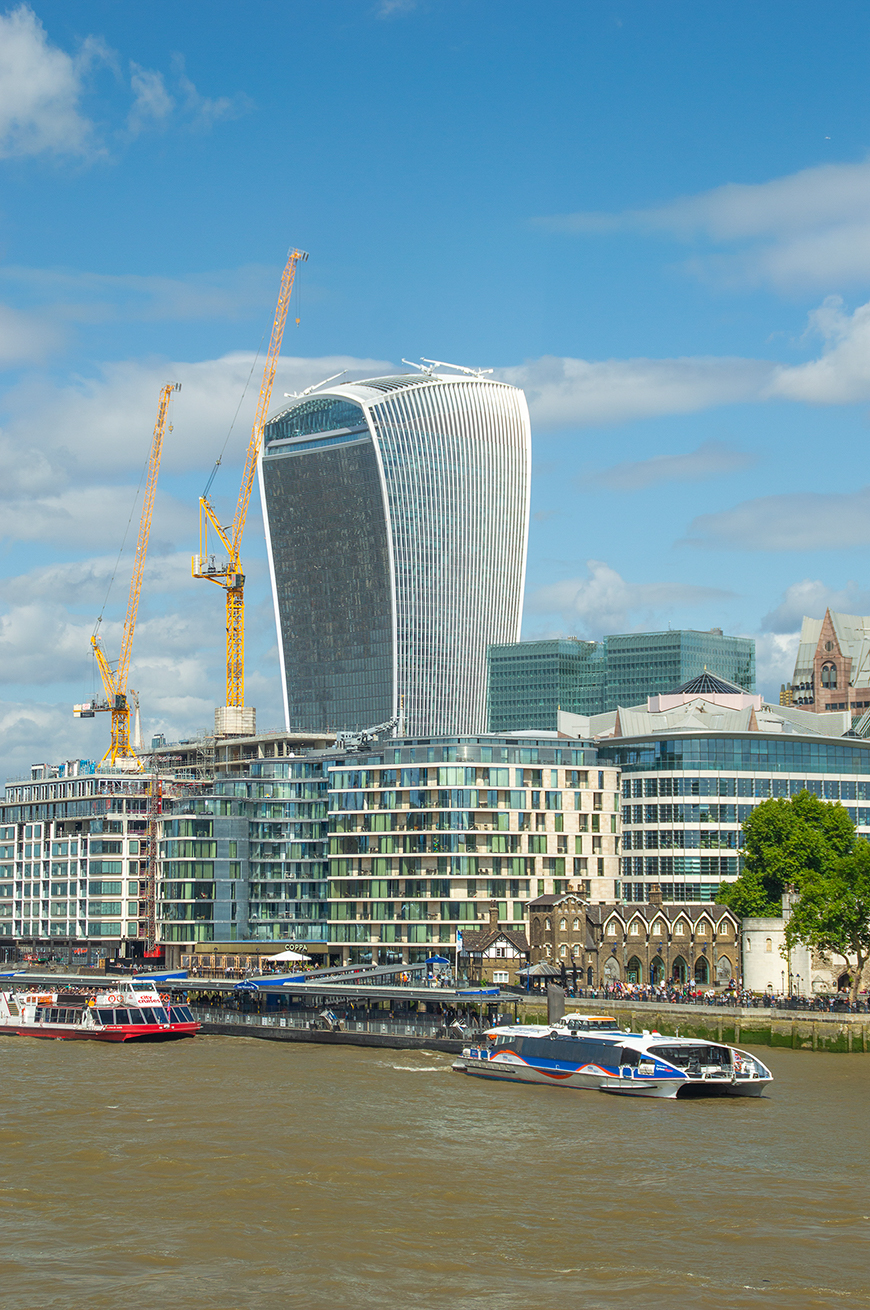 The image depicts a modern cityscape with a prominent, uniquely designed skyscraper, known as The Walkie Talkie, under construction. The scene includes several cranes, a river with boats, and a mix of contemporary and historic architecture along the waterfront.