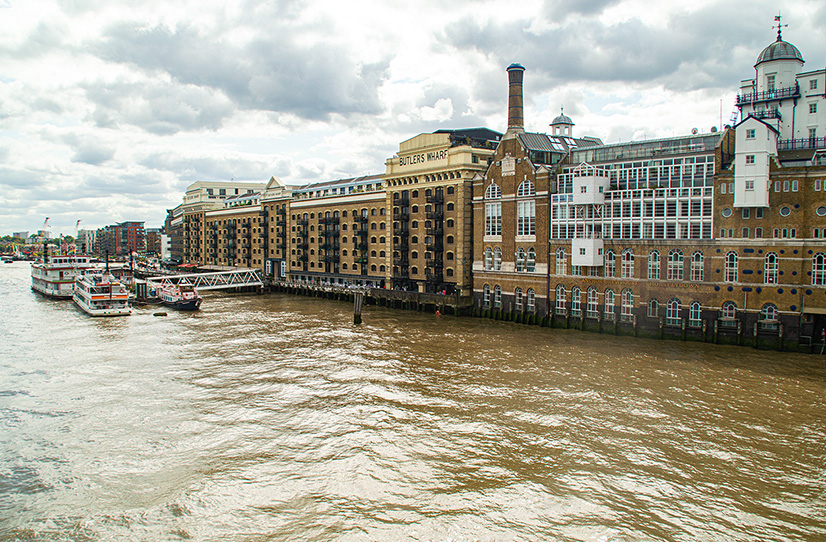 The image depicts a riverside scene with historic buildings along the bank. The buildings have a distinctive architectural style with large windows and brick facades. The river has several boats docked along the shore, and the sky is filled with clouds, suggesting an overcast day.