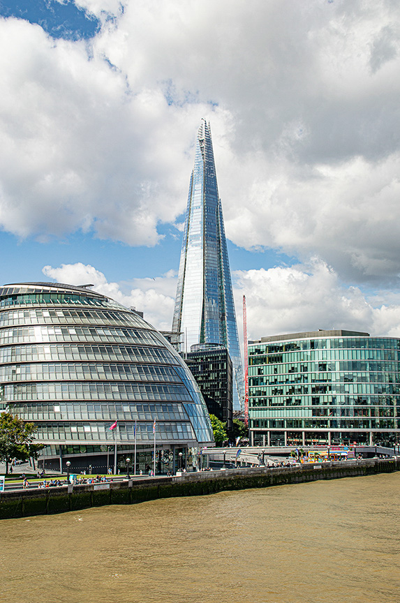 The image depicts a prominent cityscape featuring modern architecture. The Shard, a tall, pointed skyscraper, dominates the skyline. Adjacent to it is the City Hall, recognizable by its distinctive, curved shape. The scene is set along a river, with various other buildings and a partly cloudy sky in the background.