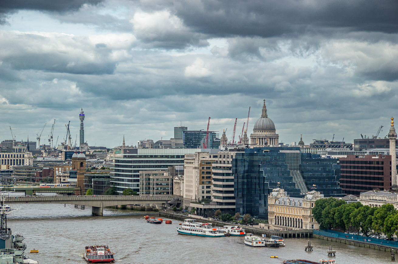 The image depicts a cityscape featuring a river with several boats, a bridge, and a variety of buildings including modern skyscrapers and historical structures. Notable landmarks include a large dome-shaped building and a tall tower with a distinctive design. The sky is overcast with dark clouds, suggesting impending rain. The scene captures a blend of architectural styles and urban activity.
