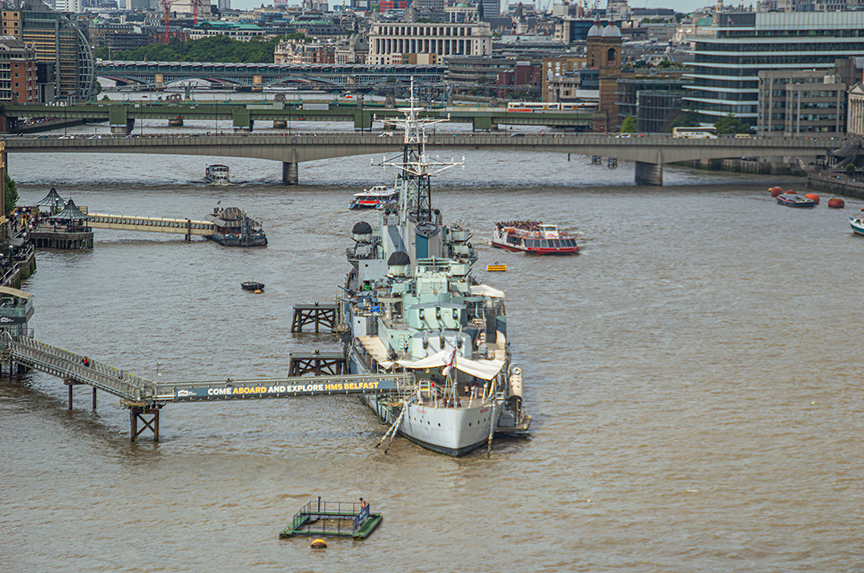 The image shows a historic naval ship docked on the River Thames in London. The ship has a sign inviting visitors to 'Come Aboard and Explore.' Surrounding the ship are various other boats and watercraft, with a bridge and cityscape in the background. The scene captures a bustling river environment with multiple vessels and urban structures.