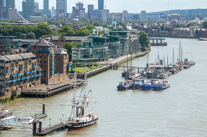The image depicts a waterfront scene with a mix of modern and historic buildings along the riverbank. Several boats and yachts are docked along the shore, and a large sailboat is seen in the foreground. The background features a city skyline with tall buildings and cranes, indicating ongoing construction and development. The area appears to be a blend of residential, commercial, and recreational spaces, with green spaces and trees interspersed among the buildings.