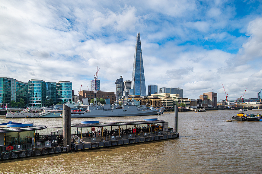 A wide view of the River Thames in London features modern buildings, including the Shard, and several ships docked along the waterway. A busy pier with people is in the foreground.