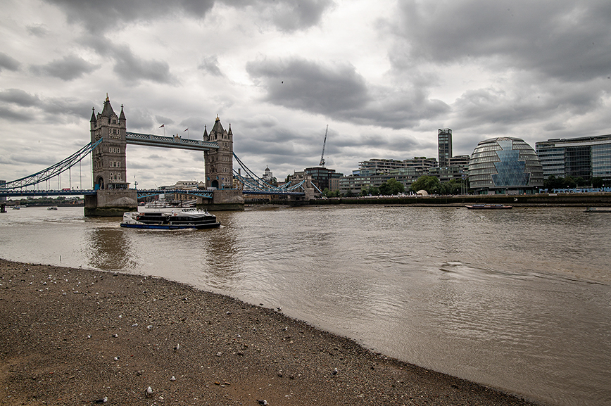 The scene looks across the River Thames on an overcast day, with Tower Bridge anchoring the left side of the composition — its grey stone towers and blue suspension elements standing out against the dull sky. A multi-deck passenger ferry moves along the brown, rippling water, while the pebble-strewn riverbank in the foreground hosts a few scattered gulls. To the right, London’s skyline stretches across the horizon: the rounded glass form of City Hall, clusters of modern office buildings, and cranes signalling ongoing development. The muted palette of greys, browns, and soft blues reinforces the somber atmosphere, while the river leads the eye from the near shore toward the historic bridge and the contemporary city beyond.