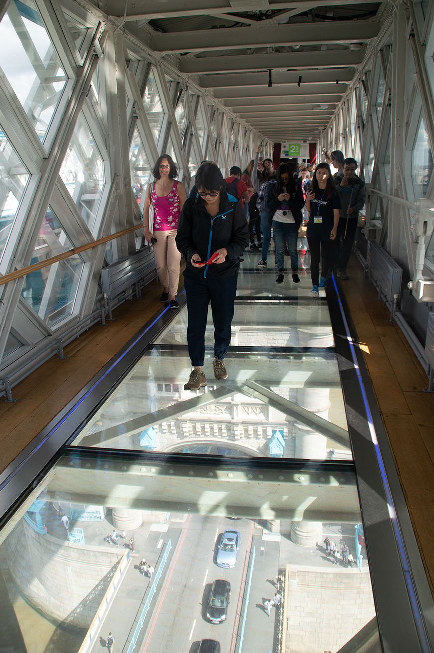 People walk along a transparent walkway with a glass floor, offering a view of traffic and pedestrians below. The walkway is enclosed by large glass windows.