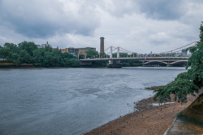 The image depicts a river with a suspension bridge crossing over it. On the left side of the river, there is a park-like area with trees and greenery. On the right side, there is a paved walkway. In the background, there are buildings and a prominent chimney structure. The sky is overcast, suggesting it might be a cloudy day.